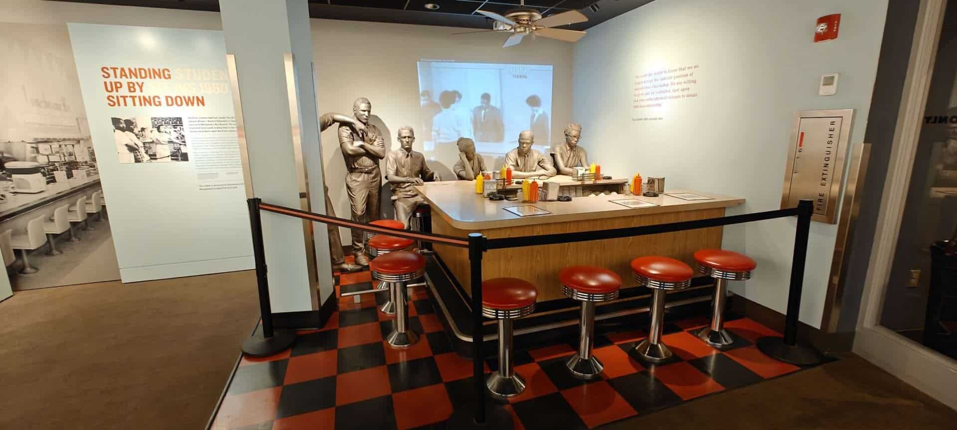 A detailed photo of a museum display featuring life-sized statues of diners, vintage stools, colorful condiments, and an informative background about vintage American diners and their cultural significance.