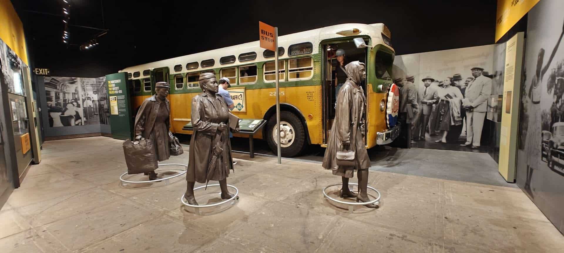 A vintage bus exhibit depicting segregation era history at a museum, featuring life-size statues of African American women with luggage, accompanied by historical photographs. The display highlights civil rights history and transportation segregation.
