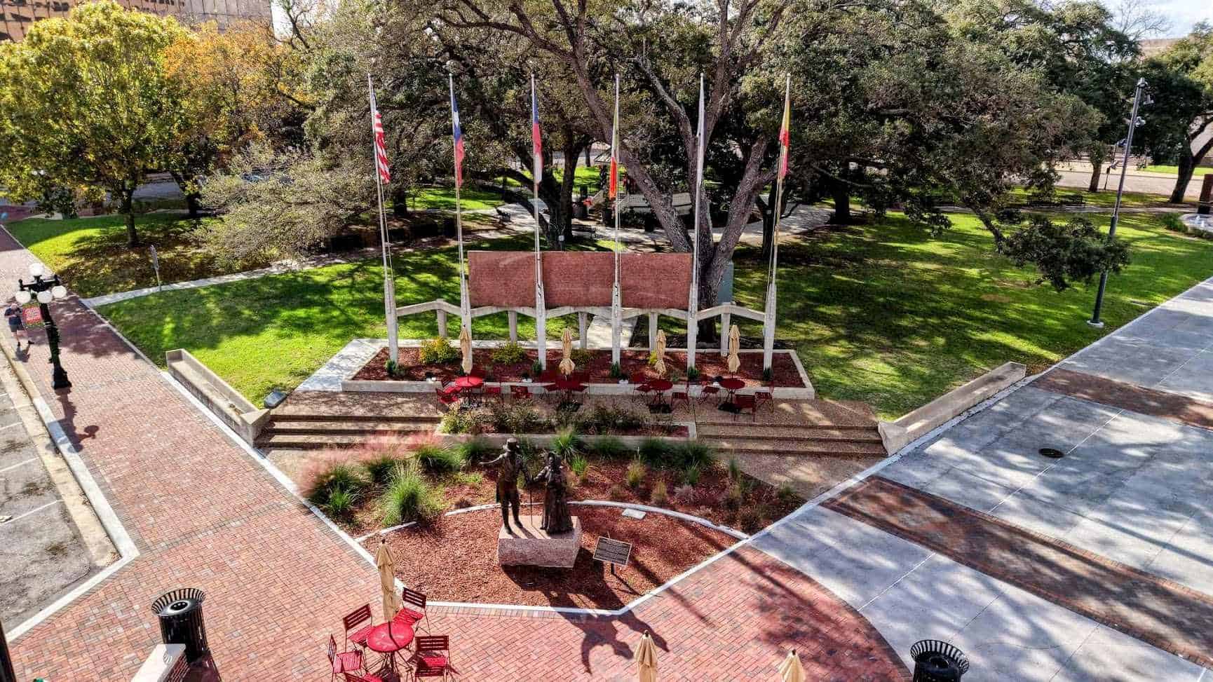 Six flagpoles and a red granite memorial wall behind the bronze De Leon founders statue at DeLeon Plaza in Victoria, Texas